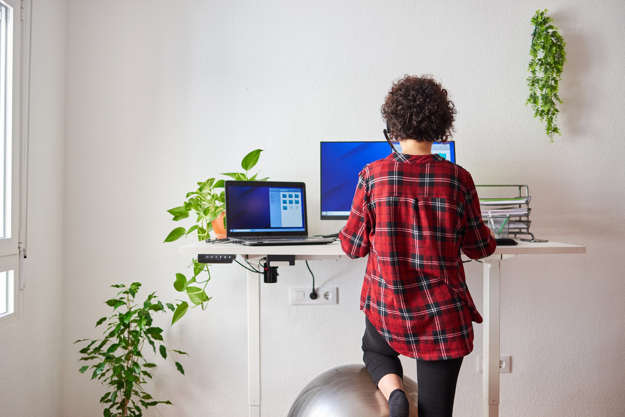A woman integrating ergonomics in her elevated home office, standing on an exercise ball while working on her computer.
